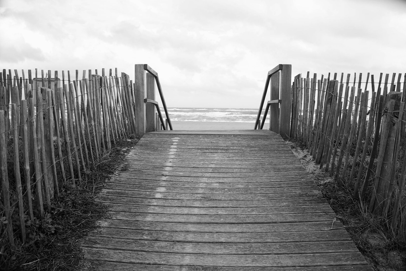 A peaceful black and white view of a wooden path leading to the ocean, framed by weathered fences.